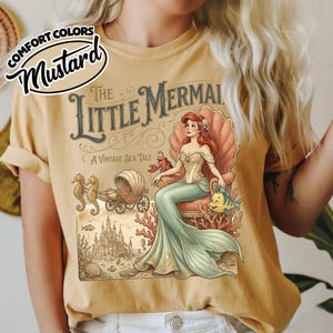 以下が含まれることがあります： マスタード色のTシャツ。ヴィンテージ風のリトルマーメイドのイラストが描かれています。デザインには、貝殻に座るアリエル、フランダー、セバスチャン、貝殻の馬車が含まれています。テキストは「The Little Mermaid: A Vintage Sea Tale」と書かれています。