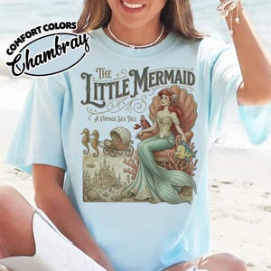 以下が含まれることがあります： 「リトル・マーメイド」のヴィンテージイラストが描かれた水色のTシャツ。貝殻の玉座に座るアリエルが描かれ、「The Little Mermaid, A Vintage Sea Tale」の文字が添えられています。シャツの左上には「Comfort Colors Chambray」の文字があります。