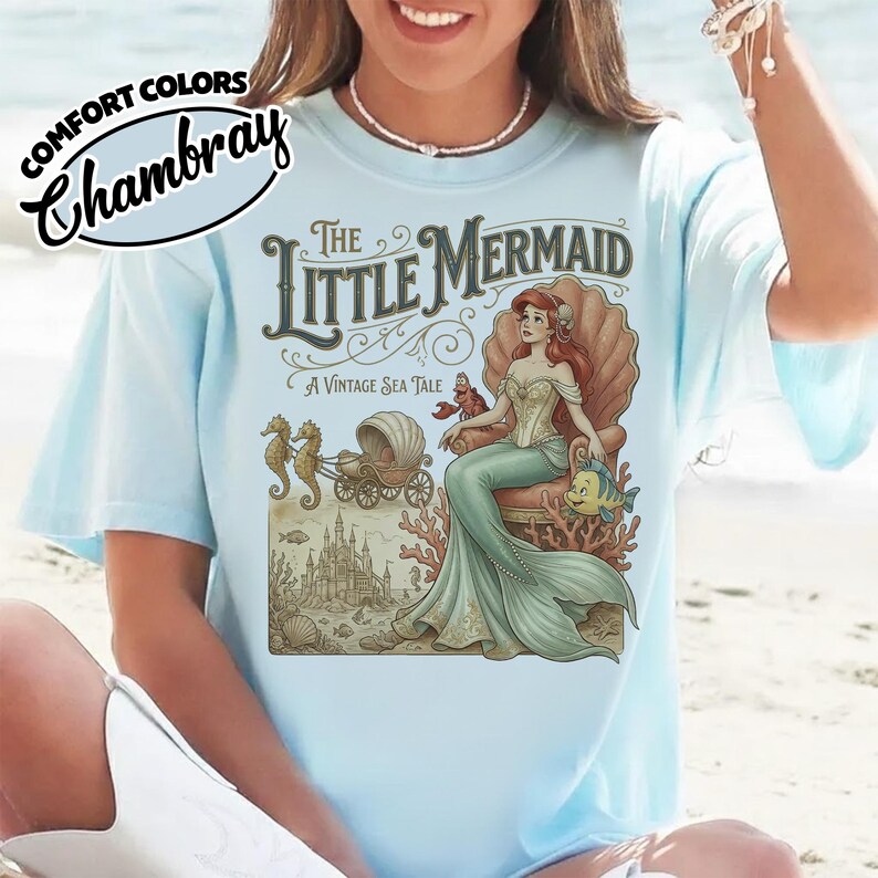 以下が含まれることがあります： 「リトル・マーメイド」のヴィンテージイラストが描かれた水色のTシャツ。貝殻の玉座に座るアリエルが描かれ、「The Little Mermaid, A Vintage Sea Tale」の文字が添えられています。シャツの左上には「Comfort Colors Chambray」の文字があります。