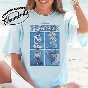 May include: Light blue t-shirt featuring the Disney movie "Frozen." The shirt displays images of Olaf, Elsa, Anna, and Sven in blue squares. The words "Disney Frozen" are printed across the top. The shirt also has the words "Comfort Colors Chambray" printed on the top left.