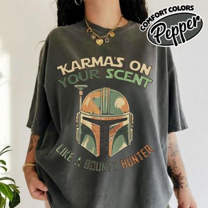 May include: Dark gray t-shirt with the text "Karma's on Your Scent" and "Like a Bounty Hunter." The shirt features a graphic of a helmet in shades of green, orange, and brown. The words "Comfort Colors Pepper" are in a circular design.