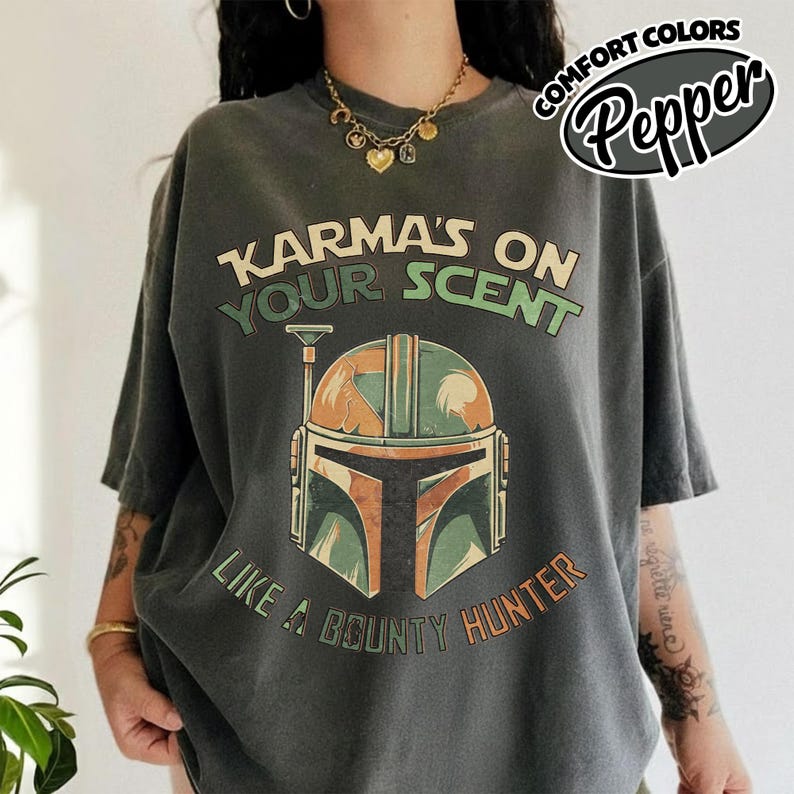 May include: Dark gray t-shirt with the text "Karma's on Your Scent" and "Like a Bounty Hunter." The shirt features a graphic of a helmet in shades of green, orange, and brown. The words "Comfort Colors Pepper" are in a circular design.
