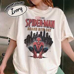 Puede incluir: Una camiseta de color marfil con un gr&aacute;fico de Spider-Man agachado frente a la silueta negra de una ciudad. El texto dice Marvel Studios Spider-Man Brand New Day. Una peque&ntilde;a etiqueta ovalada en la parte superior izquierda muestra Comfort Colors Ivory.