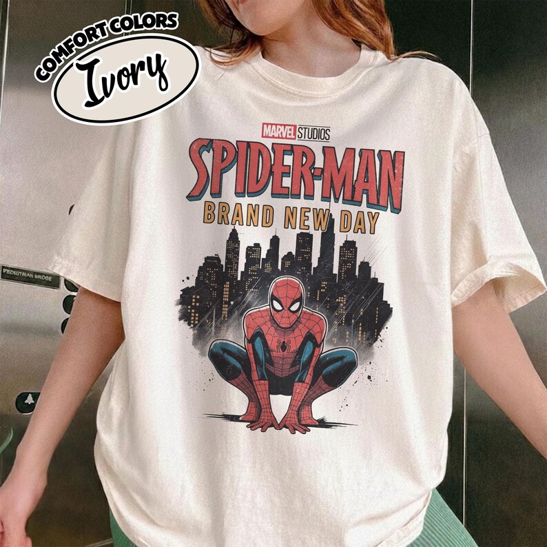 Puede incluir: Una camiseta de color marfil con un gr&aacute;fico de Spider-Man agachado frente a la silueta negra de una ciudad. El texto dice Marvel Studios Spider-Man Brand New Day. Una peque&ntilde;a etiqueta ovalada en la parte superior izquierda muestra Comfort Colors Ivory.
