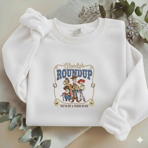 Puede incluir: Una sudadera blanca de cuello redondo con un estampado gr&aacute;fico central de Woody, Jessie, Bullseye y Stinky Pete. El texto dice Woody's ROUNDUP y YOU'VE GOT A FRIEND IN ME, enmarcado por un dise&ntilde;o de lazo con dos estrellas de sheriff abajo.