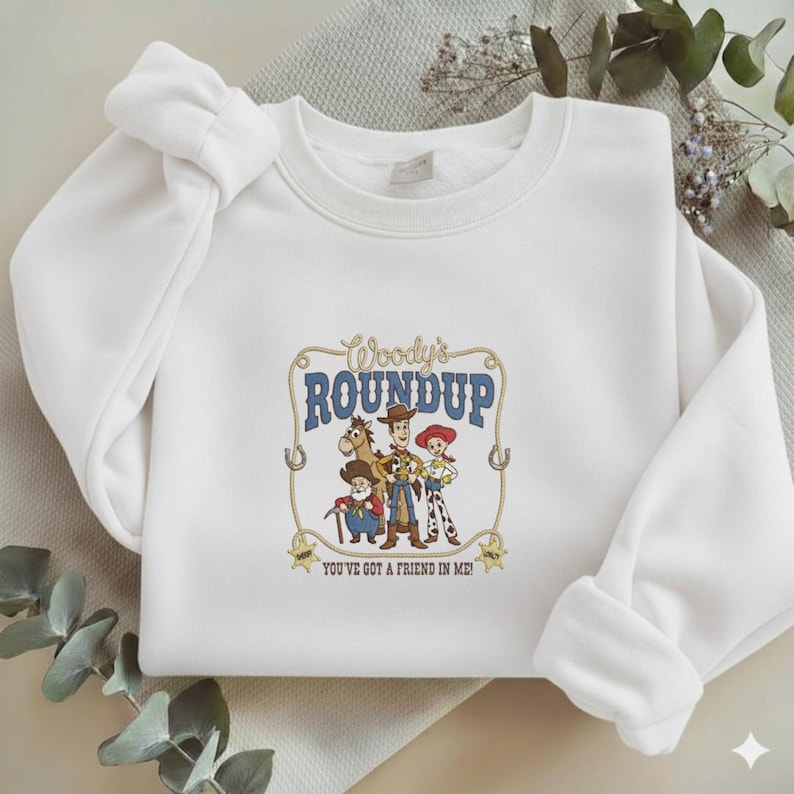 Puede incluir: Una sudadera blanca de cuello redondo con un estampado gr&aacute;fico central de Woody, Jessie, Bullseye y Stinky Pete. El texto dice Woody's ROUNDUP y YOU'VE GOT A FRIEND IN ME, enmarcado por un dise&ntilde;o de lazo con dos estrellas de sheriff abajo.