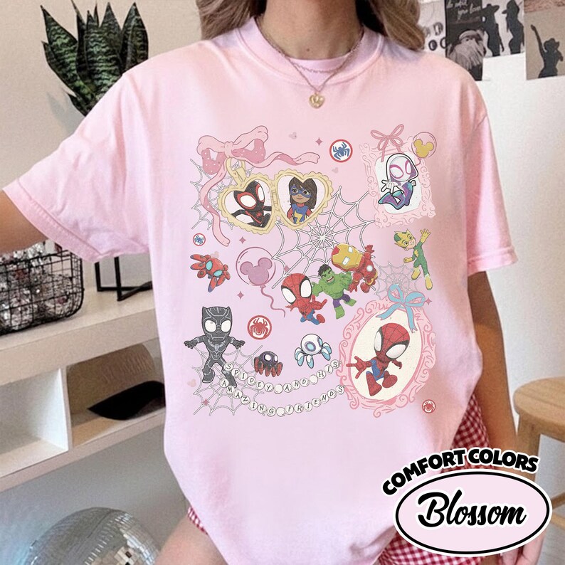 K&ouml;nnte beinhalten: Hellrosa T-Shirt mit einem farbenfrohen Aufdruck verschiedener Superhelden in dekorativen Rahmen und Spinnweben. Auf dem Shirt stehen die Worte "Amazing Spider-Friends" und "Comfort Colors Blossom".