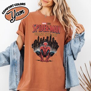 Puede incluir: Una camiseta de manga corta color &oacute;xido con un gr&aacute;fico de Spider-Man agachado frente al horizonte de una ciudad. El texto dice Marvel Studios Spider-Man Brand New Day. Un logotipo de Comfort Colors es visible en la esquina superior izquierda.