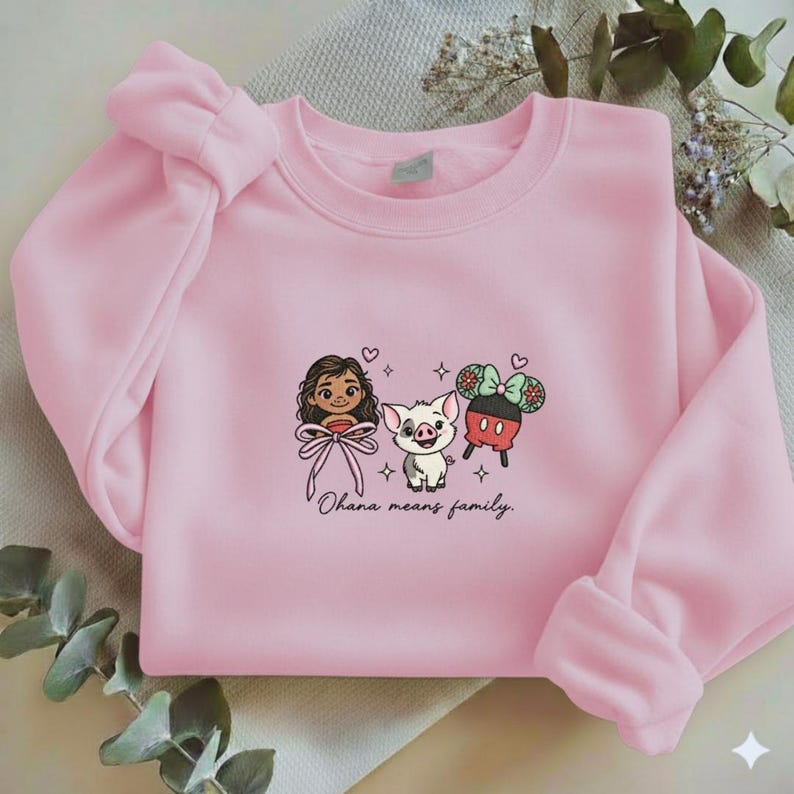 May include: A light pink crewneck sweatshirt featuring an embroidered graphic of a cartoon girl, a small pig, and a pair of mouse ears. The design includes small hearts and stars, with the cursive text "Ohana means family." printed below.