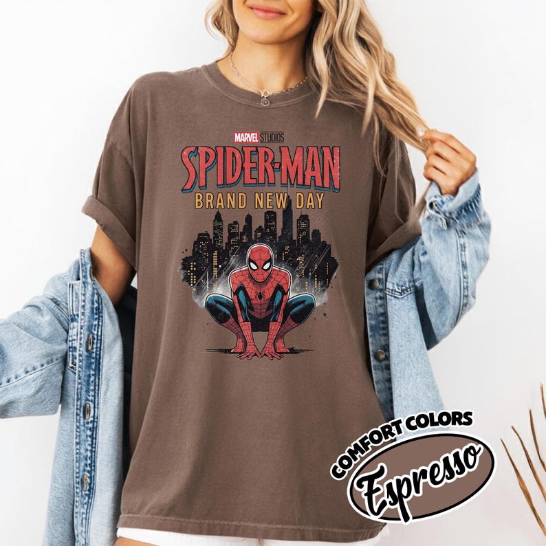 Puede incluir: Una camiseta marr&oacute;n de manga corta con un gr&aacute;fico de Spider-Man agachado frente al horizonte de una ciudad. El texto dice Marvel Studios Spider-Man Brand New Day. Una etiqueta circular en la esquina muestra el texto Comfort Colors Espresso.