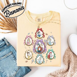 May include: A light yellow sweatshirt featuring framed portraits of animated princesses. The portraits are surrounded by decorative bows and crowns. The text "COMFORT COLORS Banana" is printed on the upper left corner.