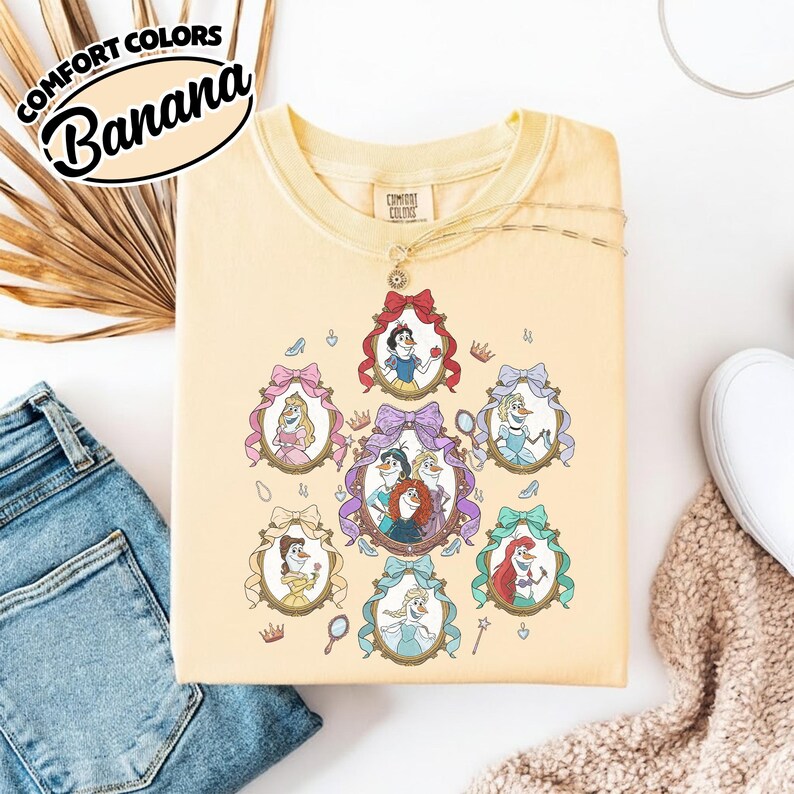 May include: A light yellow sweatshirt featuring framed portraits of animated princesses. The portraits are surrounded by decorative bows and crowns. The text "COMFORT COLORS Banana" is printed on the upper left corner.