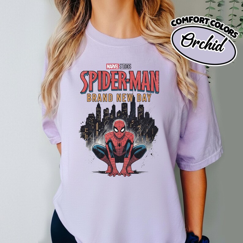 Puede incluir: Una camiseta de color morado claro con un gr&aacute;fico de Spider-Man agachado frente a la silueta negra de una ciudad. El texto dice Marvel Studios Spider-Man Brand New Day. Una peque&ntilde;a etiqueta ovalada muestra Comfort Colors Orchid.