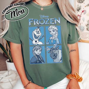May include: Green t-shirt featuring the word "FROZEN" in blue, with images of Olaf, Elsa, Anna, and Sven. The shirt has the words "COMFORT COLORS" and "Moss" in a circular design in the upper left corner.