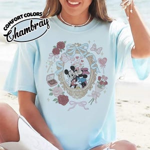 Puede incluir: Una camiseta azul claro con un gr&aacute;fico de Mickey y Minnie Mouse bes&aacute;ndose dentro de un marco dorado ornamentado. El dise&ntilde;o incluye rosas rojas y rosas, un lazo azul, una caja de bombones con la etiqueta Be Mine y corazones peque&ntilde;os en la tela.
