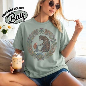 May include: Sage green t-shirt with a circular illustration of a bear and a girl holding hands, surrounded by floral and leaf designs. The shirt has the text "COMFORT COLORS Bay" in a circular design.