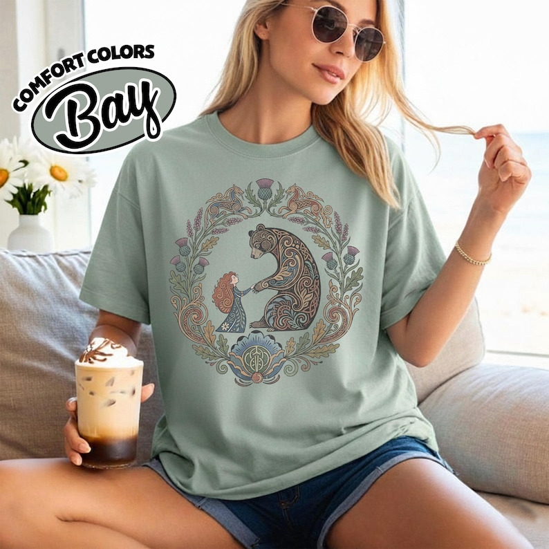 May include: Sage green t-shirt with a circular illustration of a bear and a girl holding hands, surrounded by floral and leaf designs. The shirt has the text "COMFORT COLORS Bay" in a circular design.
