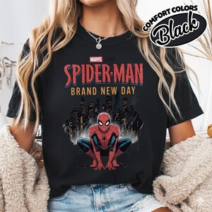 Puede incluir: Una camiseta negra con un gr&aacute;fico de Spider-Man agachado frente al horizonte de una ciudad. El texto dice Marvel Studios Spider-Man Brand New Day. Una insignia circular en la esquina superior derecha muestra el texto Comfort Colors Black.