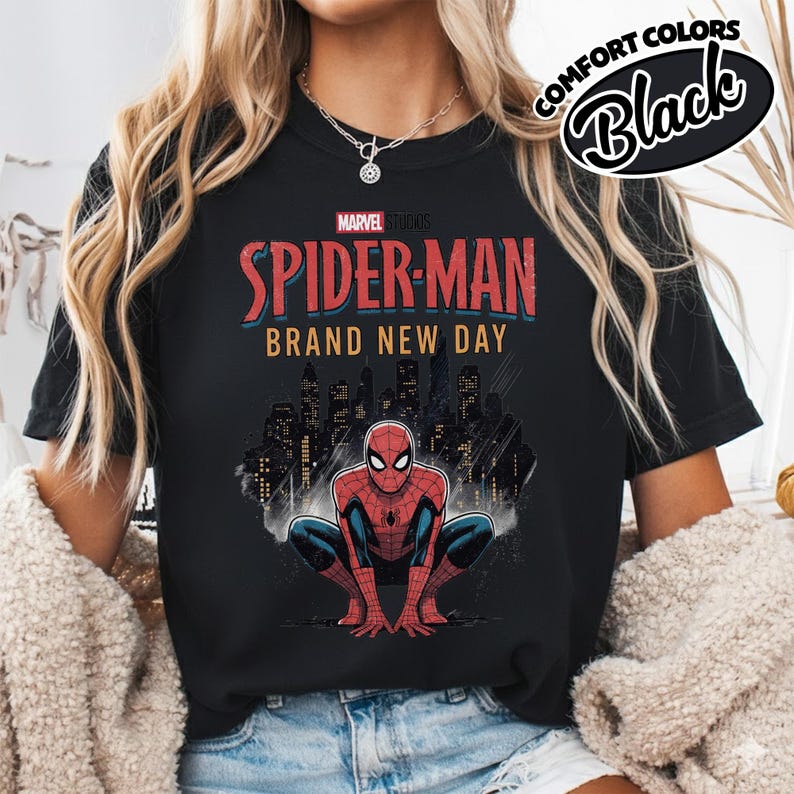 Puede incluir: Una camiseta negra con un gr&aacute;fico de Spider-Man agachado frente al horizonte de una ciudad. El texto dice Marvel Studios Spider-Man Brand New Day. Una insignia circular en la esquina superior derecha muestra el texto Comfort Colors Black.