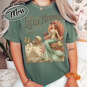 以下が含まれることがあります： モスグリーンのTシャツ。映画「リトル・マーメイド」の詳細なイラストが描かれています。デザインには、貝殻に座る人魚、城、タツノオトシゴ、その他の海の生き物が含まれています。「The Little Mermaid」の文字が装飾的なフォントで表示されています。