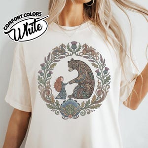 May include: Cream-colored t-shirt with a circular illustration featuring a bear and a girl holding hands, surrounded by floral and thistle elements. The shirt has the words "COMFORT COLORS White" in a circular design.