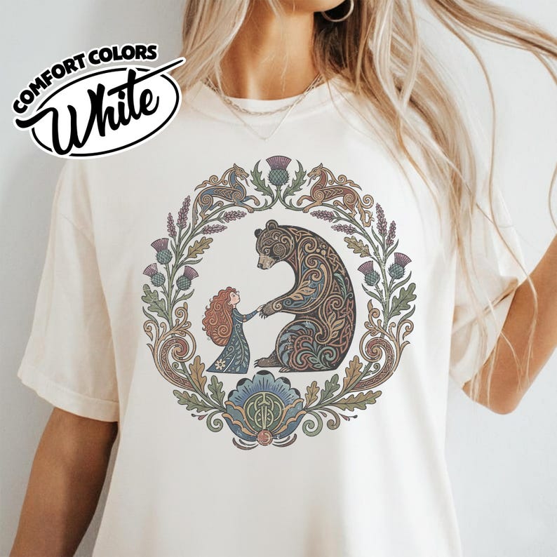May include: Cream-colored t-shirt with a circular illustration featuring a bear and a girl holding hands, surrounded by floral and thistle elements. The shirt has the words "COMFORT COLORS White" in a circular design.