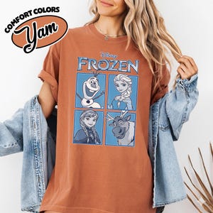 May include: Rust-colored t-shirt featuring the Disney movie "Frozen." The shirt displays four character portraits within blue squares, with the word "FROZEN" above. The top left corner has the text "COMFORT COLORS" and the word "Yam" in a stylized font.