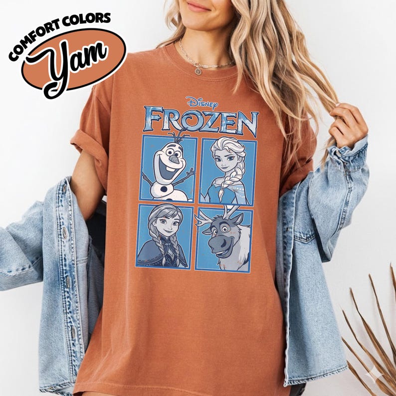May include: Rust-colored t-shirt featuring the Disney movie "Frozen." The shirt displays four character portraits within blue squares, with the word "FROZEN" above. The top left corner has the text "COMFORT COLORS" and the word "Yam" in a stylized font.