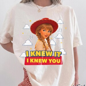 May include: An ivory short-sleeve t-shirt featuring a graphic of a character with braided hair wearing a red cowboy hat. The design includes white clouds, yellow stars, musical notes, and the text I KNEW IT I KNEW YOU in yellow and red.