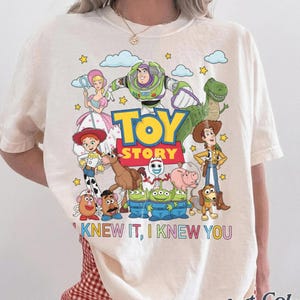 May include: An ivory short-sleeve t-shirt featuring a colorful graphic of Toy Story characters including Buzz Lightyear, Woody, Jessie, and others. The text "TOY STORY" is printed in the center, with "I KNEW IT, I KNEW YOU" written below in colorful letters.