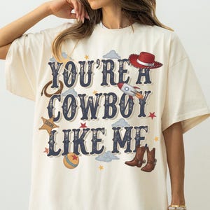 May include: An off-white short-sleeve t-shirt featuring the dark blue text "YOU'RE A COWBOY LIKE ME" surrounded by graphic illustrations of a red cowboy hat, a rocket, a lasso, a sheriff star, a colorful ball, brown cowboy boots, and small stars.