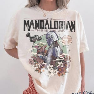 Może przedstawiać: Kremowa koszulka z grafiką The Mandalorian i Grogu otoczonych kwiatami. Tekst "THE MANDALORIAN - THIS IS THE WAY" jest wydrukowany nad obrazem. Koszulka jest marki Comfort Colors.