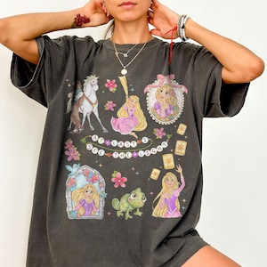 May include: Dark gray t-shirt featuring a colorful graphic of a princess with long blonde hair, a horse, and a frog. The text "AT LAST I SEE THE LIGHT" is also on the shirt. The design includes flowers and floating lanterns.