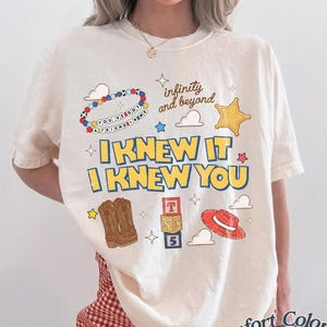 May include: An ivory short-sleeve t-shirt featuring a graphic print with the text I KNEW IT I KNEW YOU, infinity and beyond, and YOU'VE GOT A FRIEND IN ME. The design includes a beaded bracelet, a yellow star, cowboy boots, blocks, and a red hat.