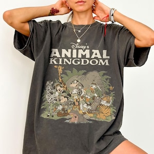 Puede incluir: Camiseta gris oscuro con el texto "Disney's Animal Kingdom" sobre un gr&aacute;fico colorido de varios animales de dibujos animados. El gr&aacute;fico incluye una cebra, una jirafa, un elefante, un tigre y varios personajes de Disney. La camiseta tiene mangas cortas y un corte holgado.