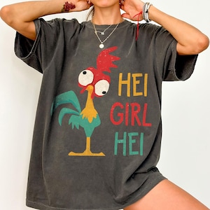 Peut inclure: T-shirt gris fonc&eacute; avec un coq de dessin anim&eacute; aux accents rouges, jaunes et turquoise. Le texte "HEI GIRL HEI" est imprim&eacute; en jaune, rouge et turquoise. Le t-shirt est ample et &agrave; manches courtes.