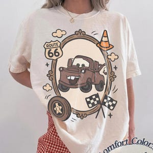 May include: Ivory-colored t-shirt featuring a cartoon image of a brown truck inside a decorative frame, with Route 66 signage, a traffic cone, and checkered flags. The shirt is labeled "Comfort Colors Ivory".