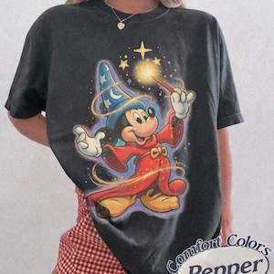 Pu&ograve; includere: T-shirt grigio scuro con un design di Topolino dei cartoni animati vestito da mago, che tiene in mano una bacchetta con effetti scintillanti. Il design include un cappello da mago blu con stelle e una veste rossa. La maglietta ha il testo "Comfort Colors Pepper" in un ovale bianco.