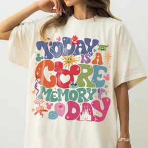 May include: Cream-coloured oversized t-shirt featuring the phrase "Today is a Core Memory Day" in colourful, retro lettering. The design includes cartoon characters and a heart-shaped Mickey Mouse head. A fun, whimsical graphic tee.