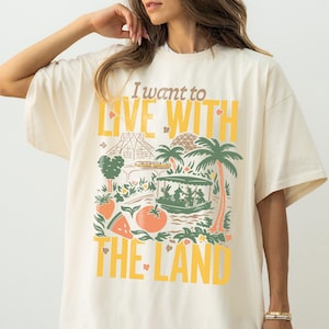 Könnte beinhalten: Cremefarbenes T-Shirt mit dem Text "I want to LIVE WITH THE LAND" in Gelb und Braun. Die Grafik zeigt ein Boot, Palmen und verschiedene Früchte und Gemüse, die ein tropisches Thema hervorrufen.