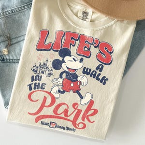 May include: Cream-coloured t-shirt featuring Mickey Mouse and the text "LIFE'S A WALK IN THE PARK" in red and blue. The design includes an illustration of Cinderella's castle and the words "Walt Disney World".