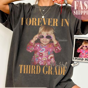 Op de afbeelding: Een antracietgrijs t-shirt met korte mouwen en een vintage-stijl afbeelding van een kind met een roze zonnebril en een shirt met bloemenpatroon. De tekst luidt FOREVER IN THIRD GRADE en bevat de handgeschreven naam Ms Hall.