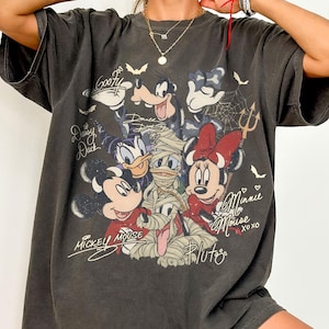 May include: Dark grey t-shirt with a Halloween-themed graphic of Disney characters. Mickey, Minnie, Donald, Daisy, Goofy, and Pluto are shown in costume. The shirt includes the text "Mickey Mouse" and "Minnie Mouse xoxo".