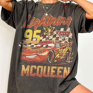 May include: Dark gray t-shirt featuring a graphic of Lightning McQueen from the Cars movie. The design includes the text "Lightning 95 McQueen" in yellow and red, with a checkered flag and atomic symbol.