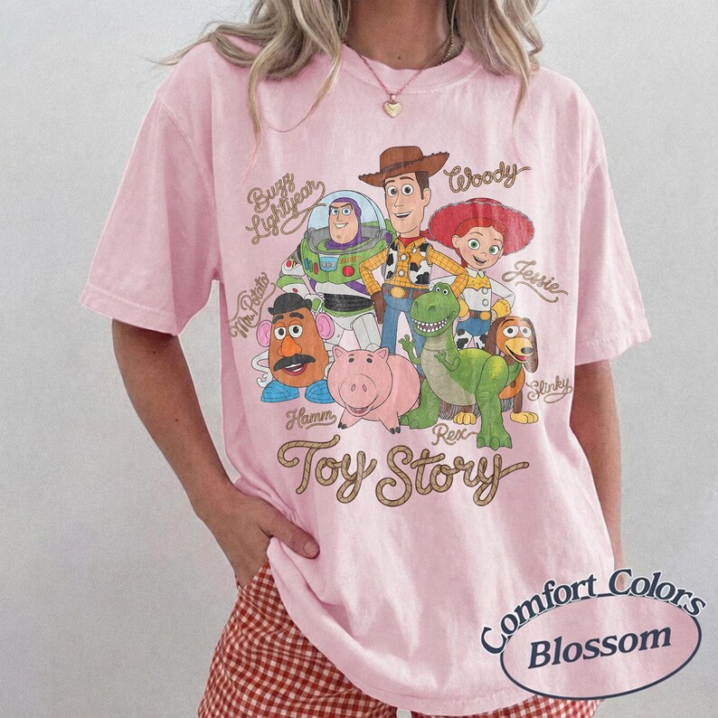 May include: A light pink short-sleeve t-shirt featuring a graphic print of Toy Story characters including Woody, Buzz Lightyear, Jessie, Mr. Potato Head, Hamm, Rex, and Slinky Dog. The text Toy Story is printed below the characters in a cursive font.