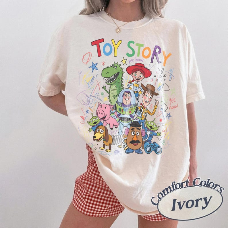 May include: Ivory-colored Comfort Colors t-shirt featuring a colorful graphic of Toy Story characters and the words "TOY STORY" in rainbow letters. The design includes Woody, Buzz Lightyear, and other beloved characters.