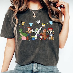 May include: Dark gray t-shirt featuring a colorful graphic of animated characters from a popular movie. The design includes characters like a dinosaur, cowboy, space ranger, and horse, with Mickey Mouse-shaped balloons above them. The shirt is a casual wear item.