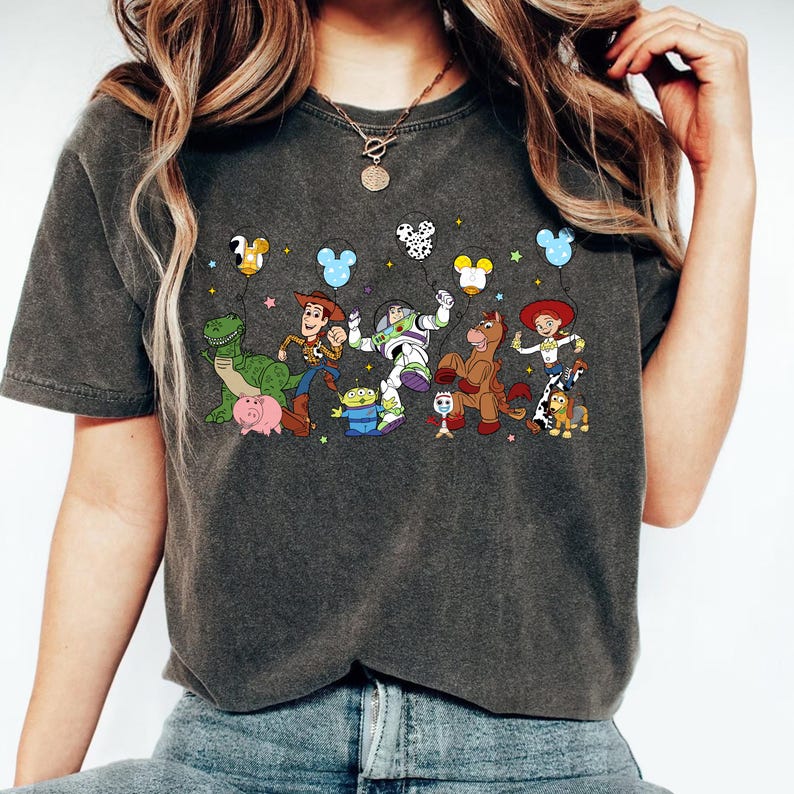 May include: Dark gray t-shirt featuring a colorful graphic of animated characters from a popular movie. The design includes characters like a dinosaur, cowboy, space ranger, and horse, with Mickey Mouse-shaped balloons above them. The shirt is a casual wear item.