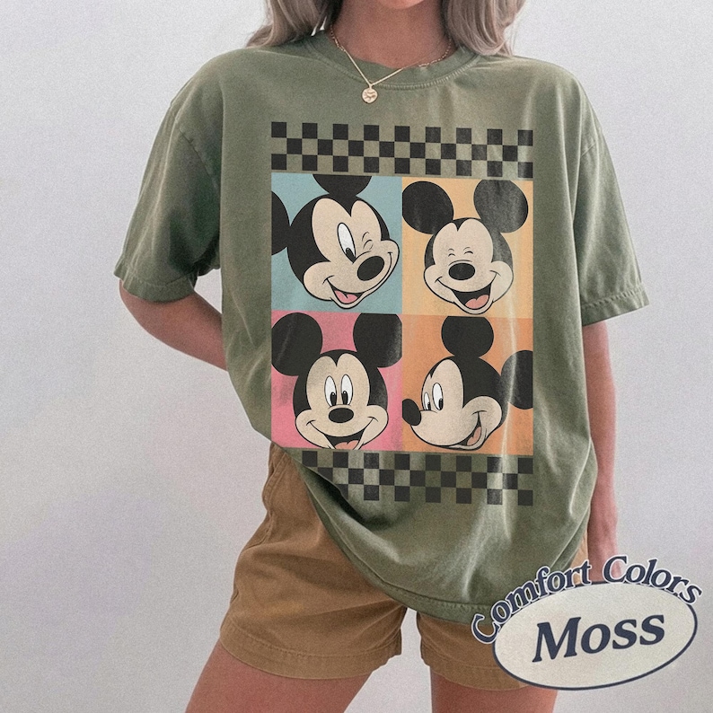 May include: Olive green t-shirt featuring four panels with Mickey Mouse's face in different expressions. The panels are in blue, orange, pink, and orange, with a black and white checkered pattern above and below. The text "Comfort Colors Moss" is printed on the bottom right.