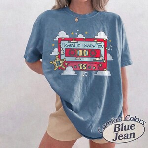 May include: A blue short-sleeve t-shirt featuring a graphic of a red cassette tape with the text "I KNEW IT, I KNEW YOU" and "TS". The design includes white clouds, yellow stars, and musical notes. The shirt has a crew neckline and a relaxed fit.